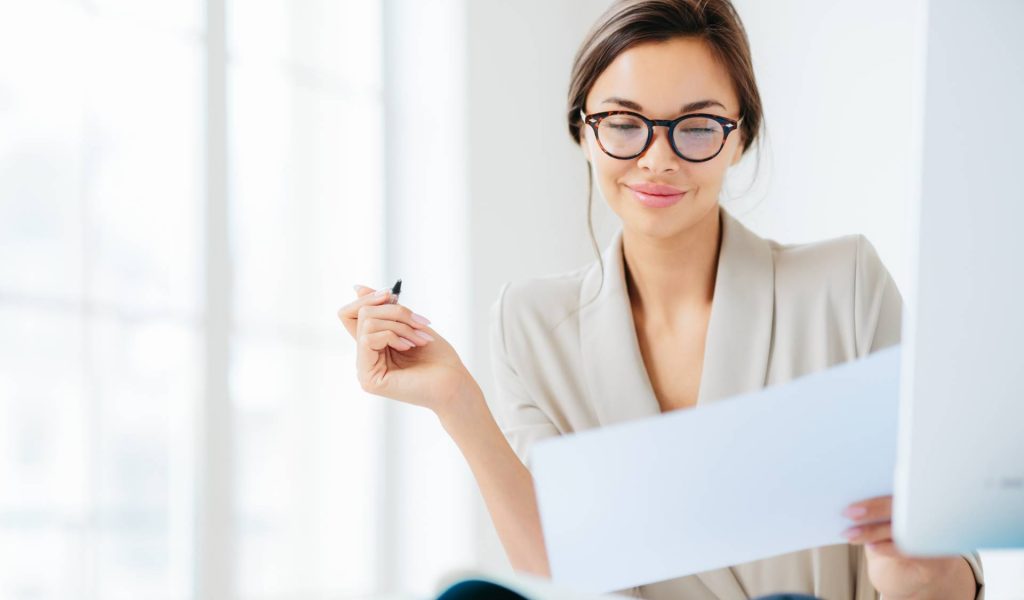 Concentrated successful businesswoman looks attentively in paper, studies terms of contract, holds pen, writes in documentations, dressed formally, poses at desktop against white spacious interior