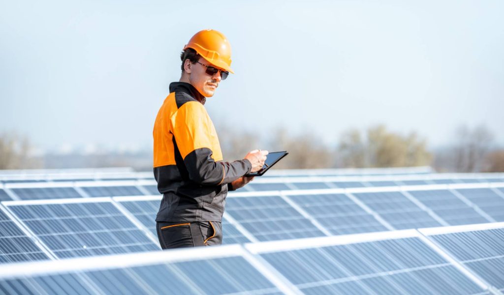 Engineer in protective workwear carrying out service of solar panels with digital tablet on a photovoltaic rooftop plant. Concept of maintenance and setup of solar power station