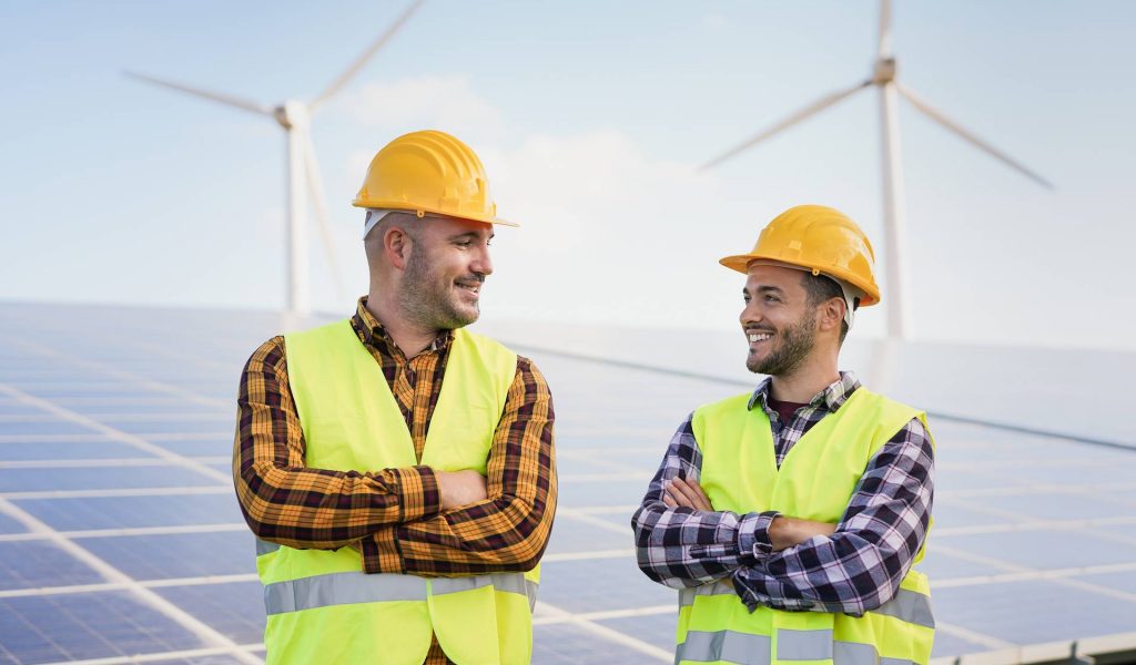 Worker men at solar power station - Solar panels with wind turbines in background - Green energy renewable concept